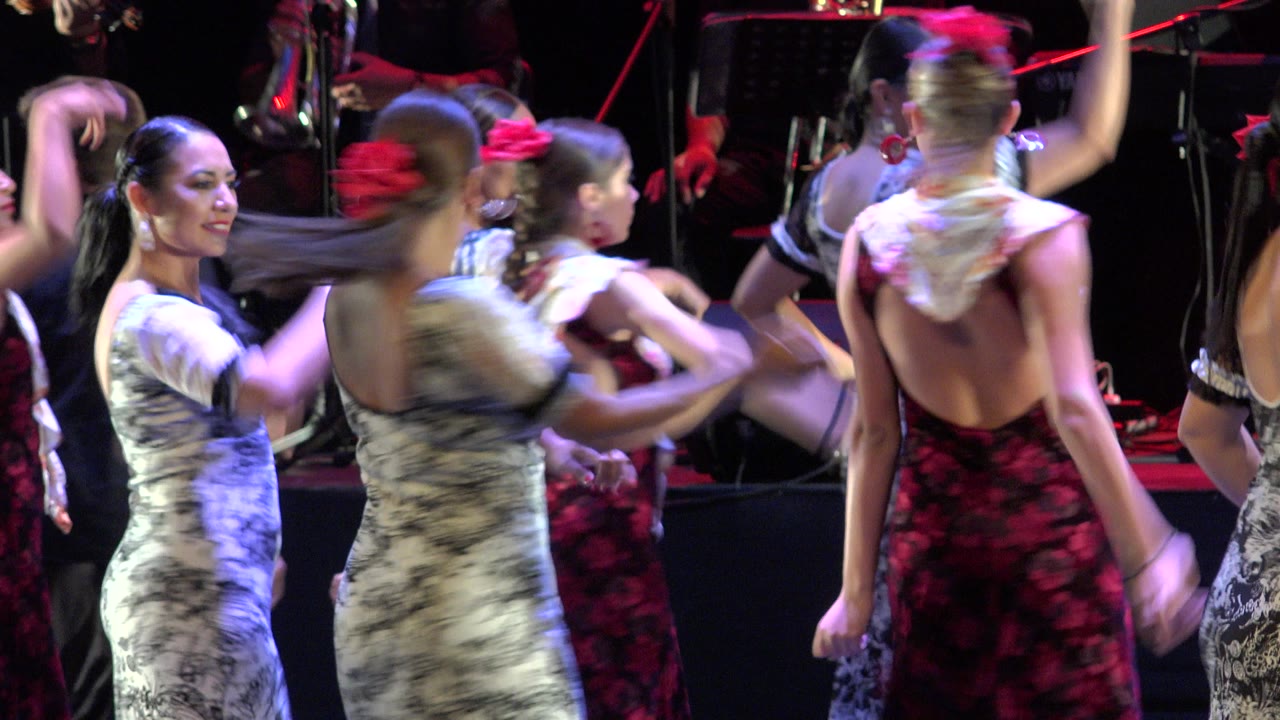 Dancers in floral dresses lift their arms during a stage routine in Cabo San Lucas, Mexico, with a violinist and other musicians visible behind them.