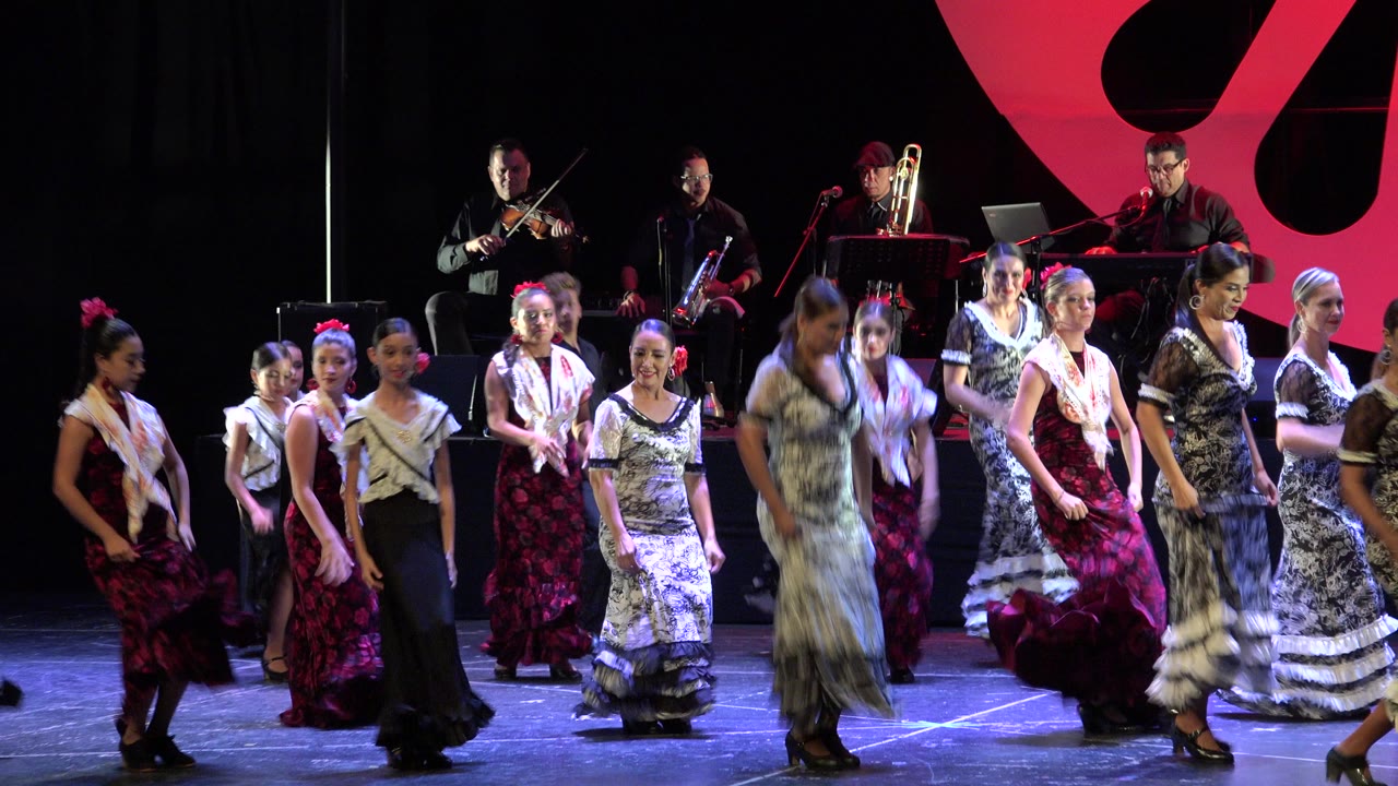 Flamenco dancers perform on a lit stage in Cabo San Lucas, Mexico, with musicians seated on a raised platform behind them at the opening moment.