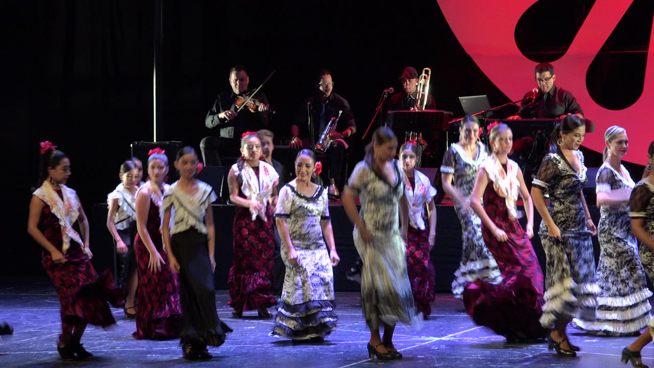 A line of flamenco dancers moves across the Cabo San Lucas stage while violin, brass, and keyboard players remain visible in the background.