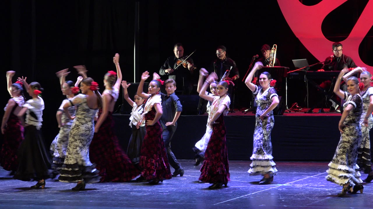 Women in full-length dresses move across the lit stage in Cabo San Lucas, Mexico, with several musicians seated and standing on the rear platform.