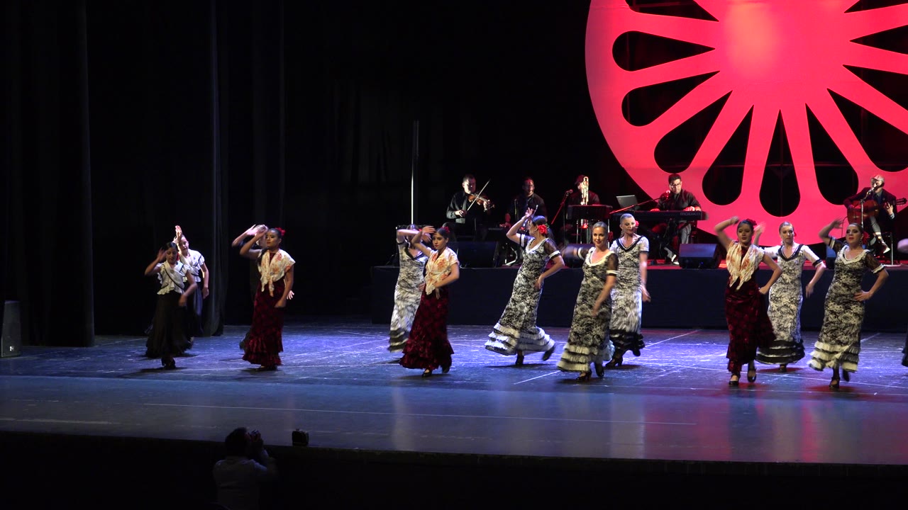 Wide view of a Cabo San Lucas stage performance with dancers spread across the lit floor, musicians on rear risers, black curtains, and a large red fan-shaped backdrop.