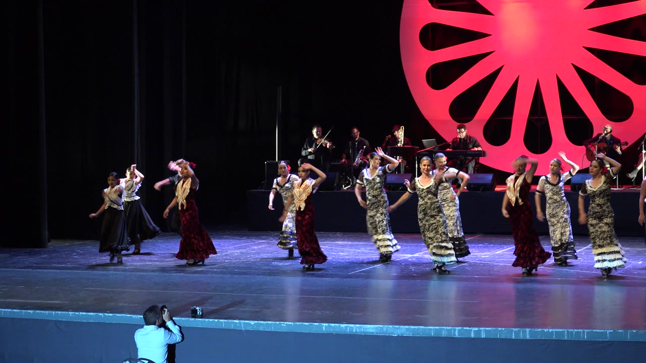 Wide Cabo San Lucas performance view with dancers across the stage, musicians behind them, and a person visible near the front of the audience filming toward the performers.