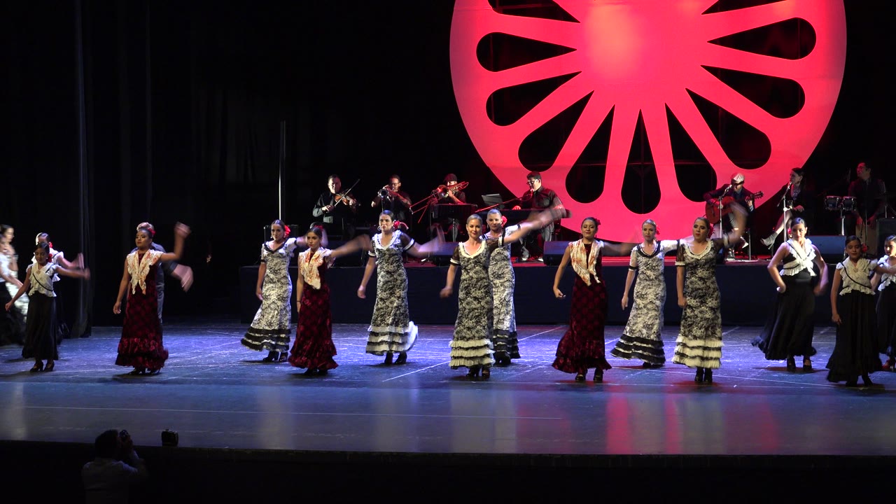 Dancers in ruffled dresses line up across a theater stage with musicians behind them.