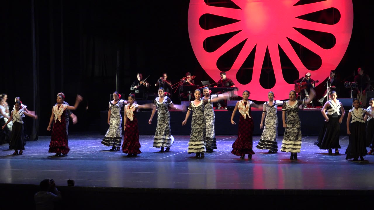 The Cabo San Lucas performance continues in a wide stage view as dancers in patterned and red dresses face forward beneath the bright red fan-shaped set piece.