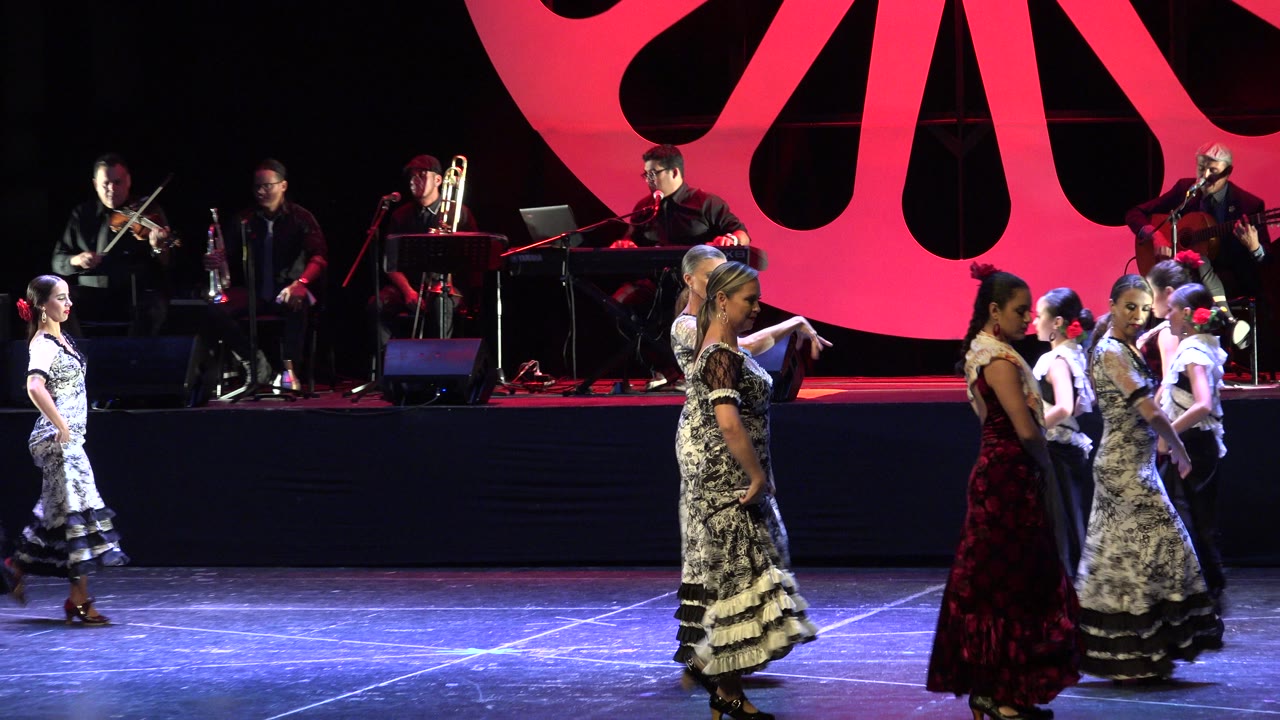 Young women in patterned dresses dance on a stage with musicians behind them.