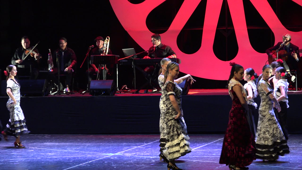One second in, the Cabo San Lucas stage remains wide, with dancers moving left to right and a live band seated against a large red and black backdrop.