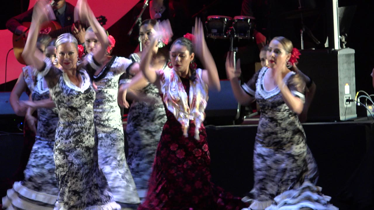 At 0:01, the dancers remain grouped across the Cabo San Lucas stage, with several arms lifted and a woman in a red and black skirt moving near the foreground.