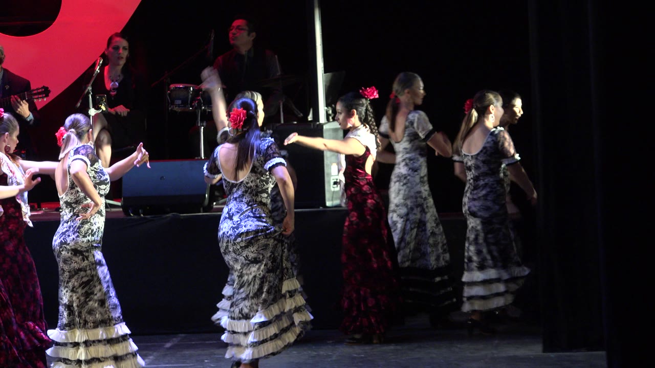 Adult women in patterned black, white, gray, and red dresses dance on a stage in Cabo San Lucas, Mexico, with red flowers in their hair and musicians visible behind them.
