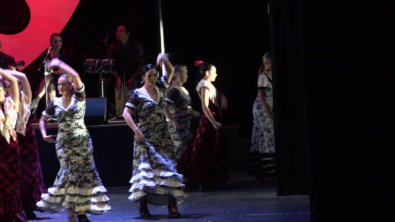 Women in ruffled dresses raise arms and move across a lit stage.