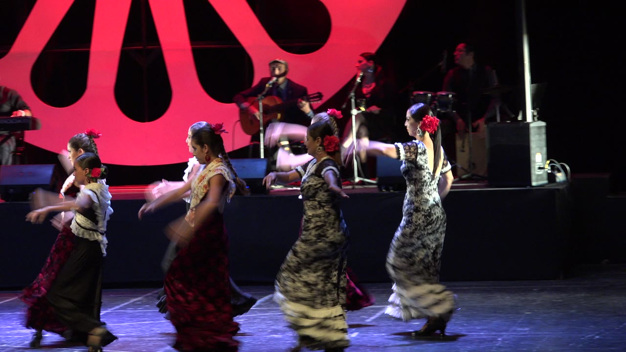 Young dancers cross a stage in front of live musicians and a red backdrop.