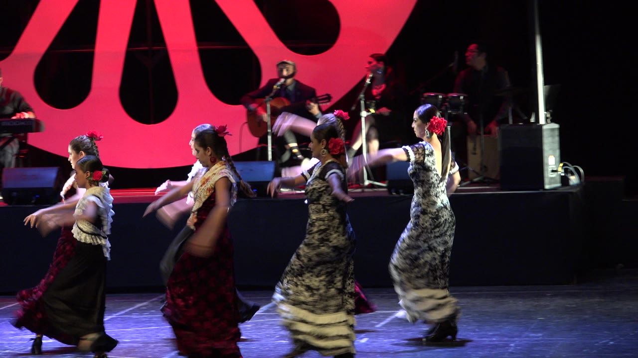 Several young dancers continue across the Cabo San Lucas stage in long red, black, and white dresses with musicians partly visible behind them.