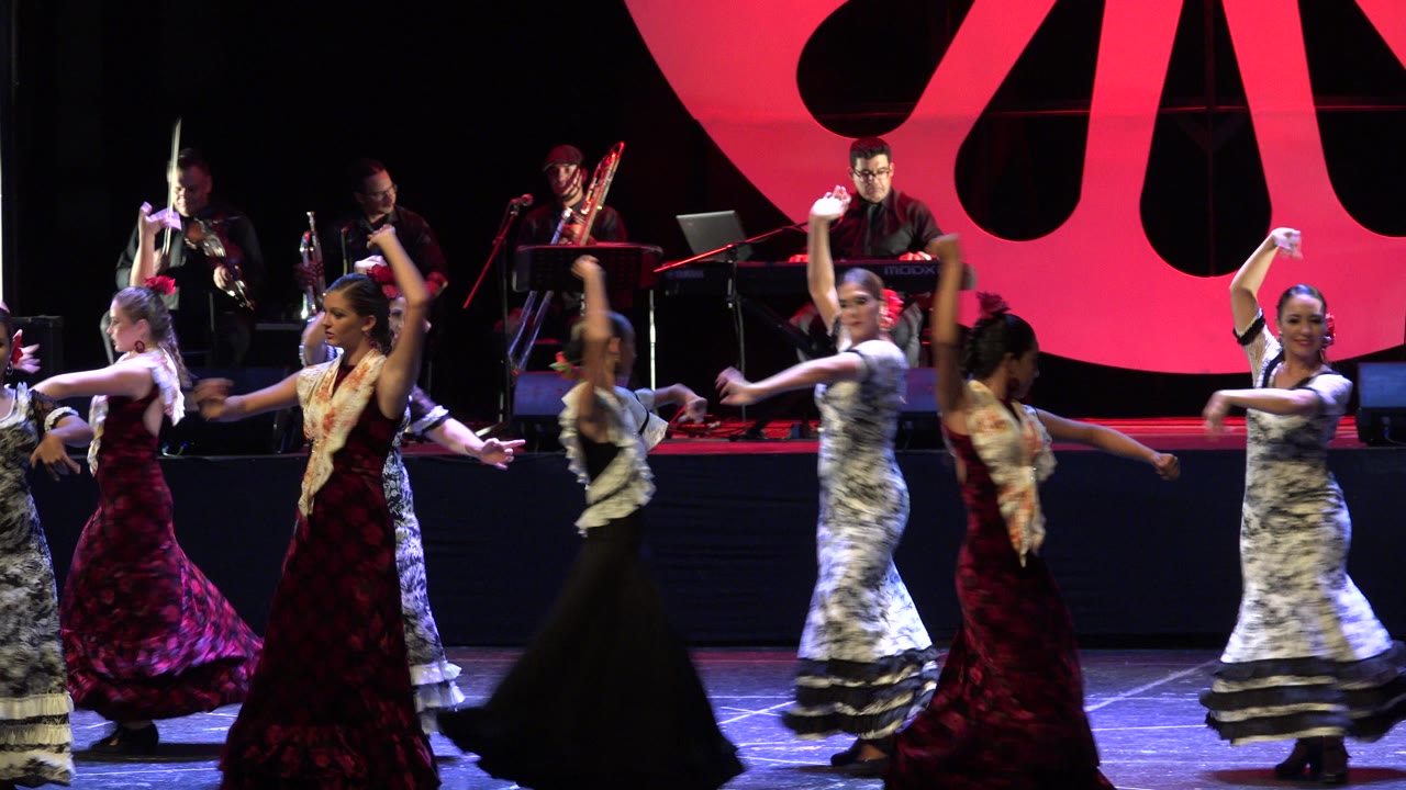 A line of dancers turns across the foreground in Cabo San Lucas, Mexico, while the band remains on the raised platform behind the routine.