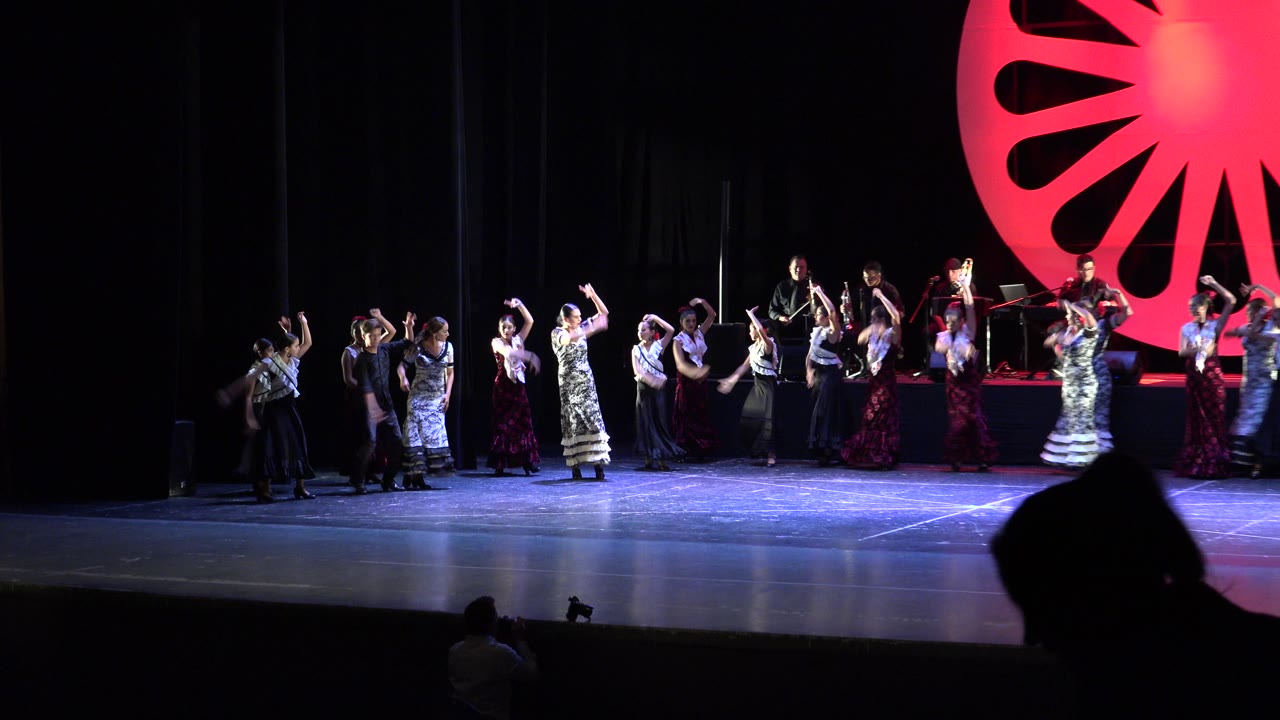 Wide audience view in Cabo San Lucas, Mexico, showing children and adult dancers spread across a dark stage while musicians sit on a rear platform under a large red circular set piece.