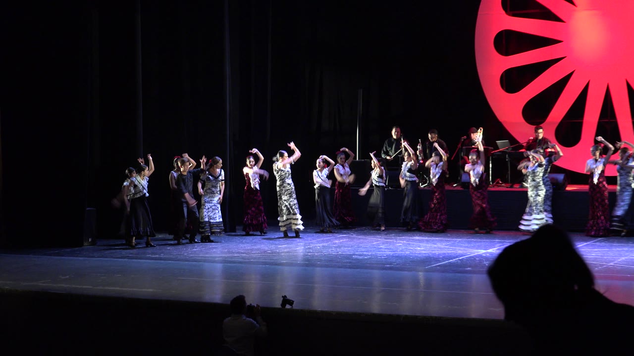 Audience-level frame in Cabo San Lucas, Mexico, with multiple young and adult performers holding raised-arm dance positions as musicians remain seated behind them on the lit stage.