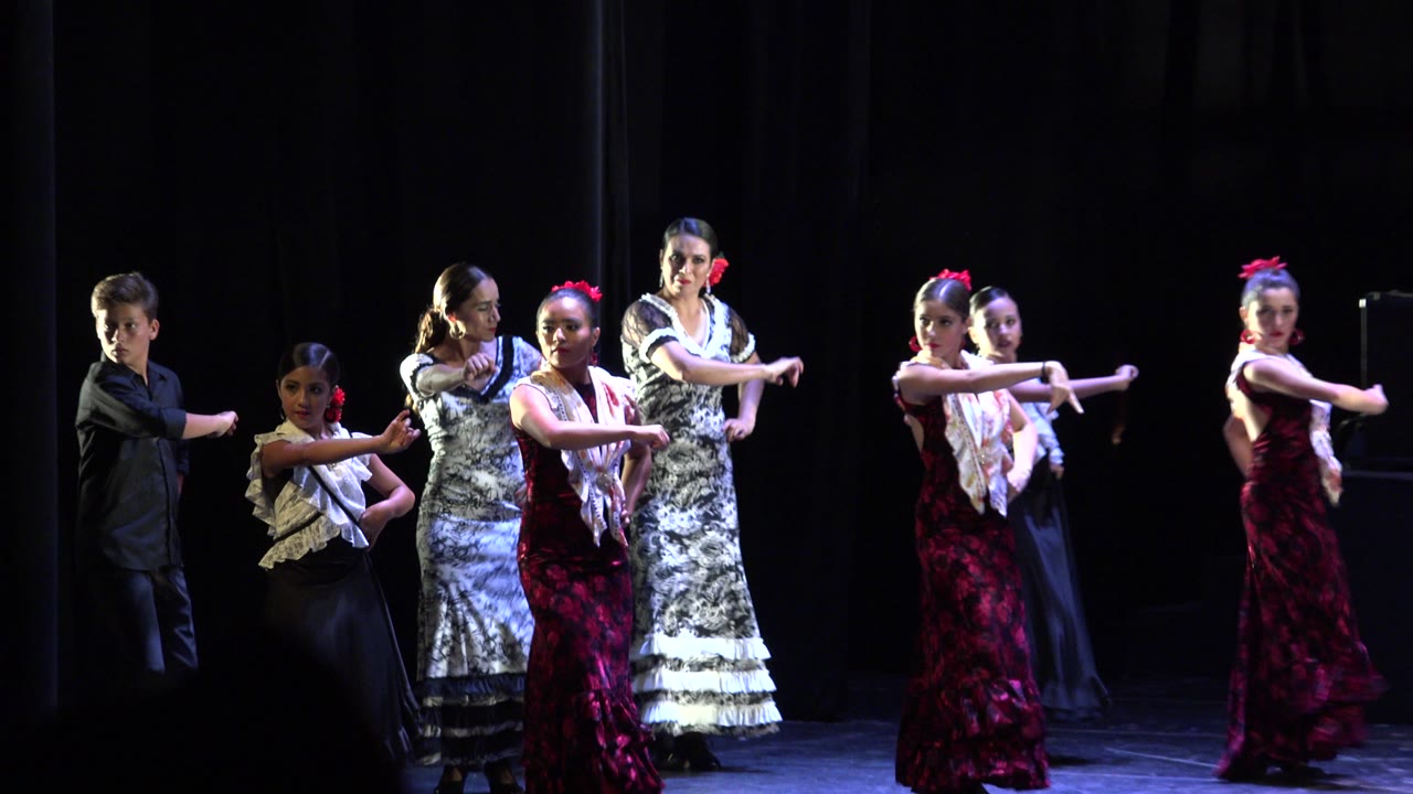 A group of performers in Cabo San Lucas remains spread across the dark stage, with several faces visible and arms held outward in a coordinated pose.