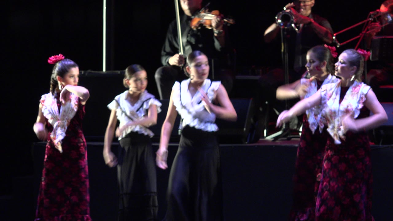 Young dancers in white and red performance outfits shift into a synchronized gesture while violin, trombone, and other musicians remain visible in the background in Cabo San Lucas, Mexico.