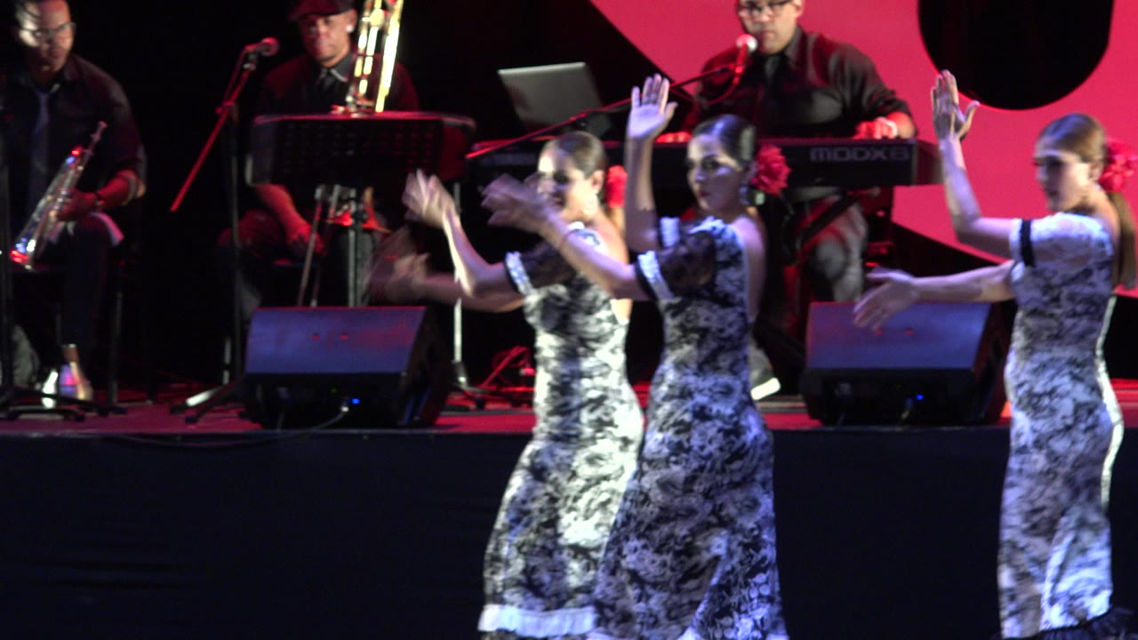Young girls in patterned dresses dance on a Cabo San Lucas stage while musicians with saxophone, trombone, keyboard, and microphones play behind them.