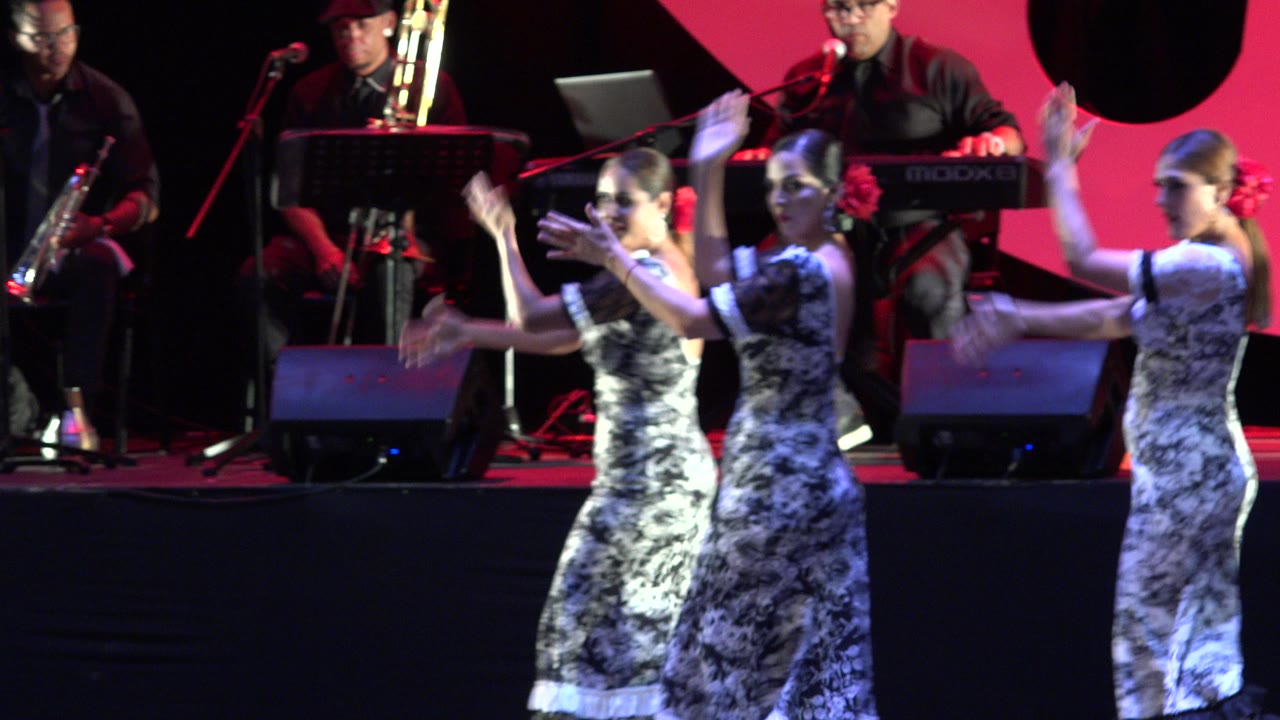 Three girls in ruffled dresses raise their arms in a synchronized dance on a Cabo San Lucas stage, with a keyboardist and brass players in the background.
