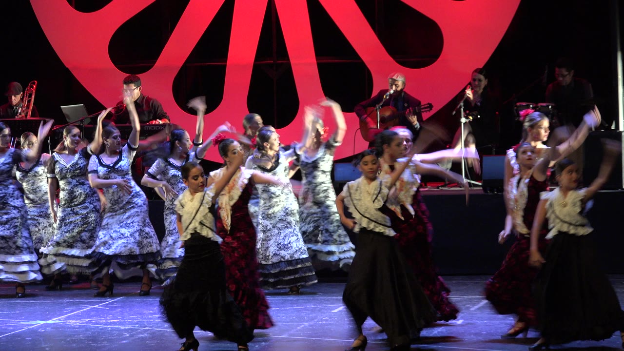 Girls dancing on a stage with musicians behind them and a large red backdrop.