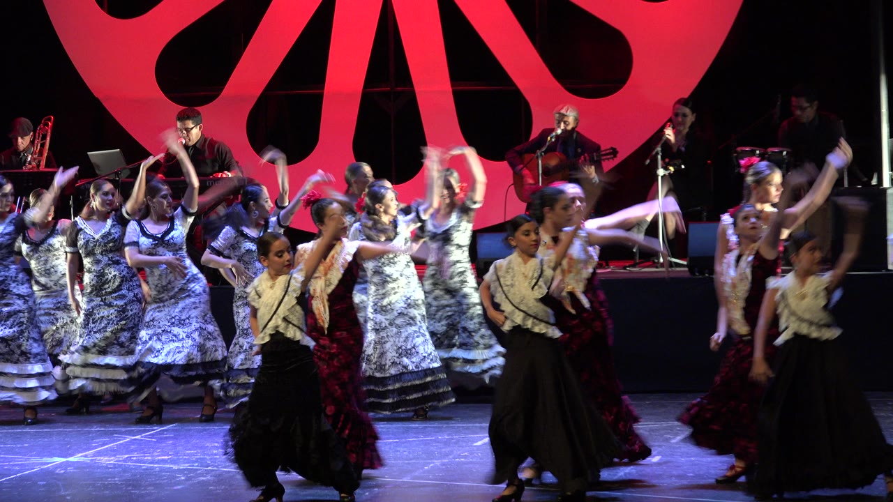 A group of girls in coordinated black, white, and red costumes dance across the Cabo San Lucas stage as musicians perform on the rear platform.