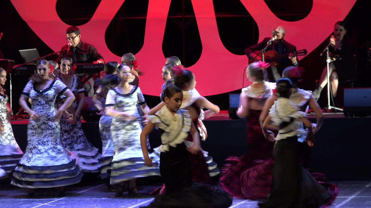 Girls in flamenco-style dresses dance on a stage with musicians behind them.