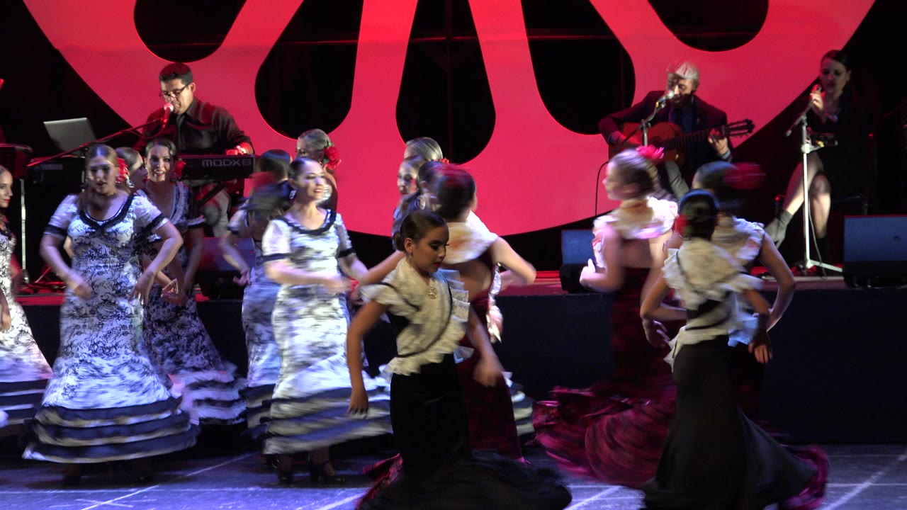 Young dancers in white, black, and red flamenco-style costumes cross the Cabo San Lucas stage as musicians remain visible in the rear.