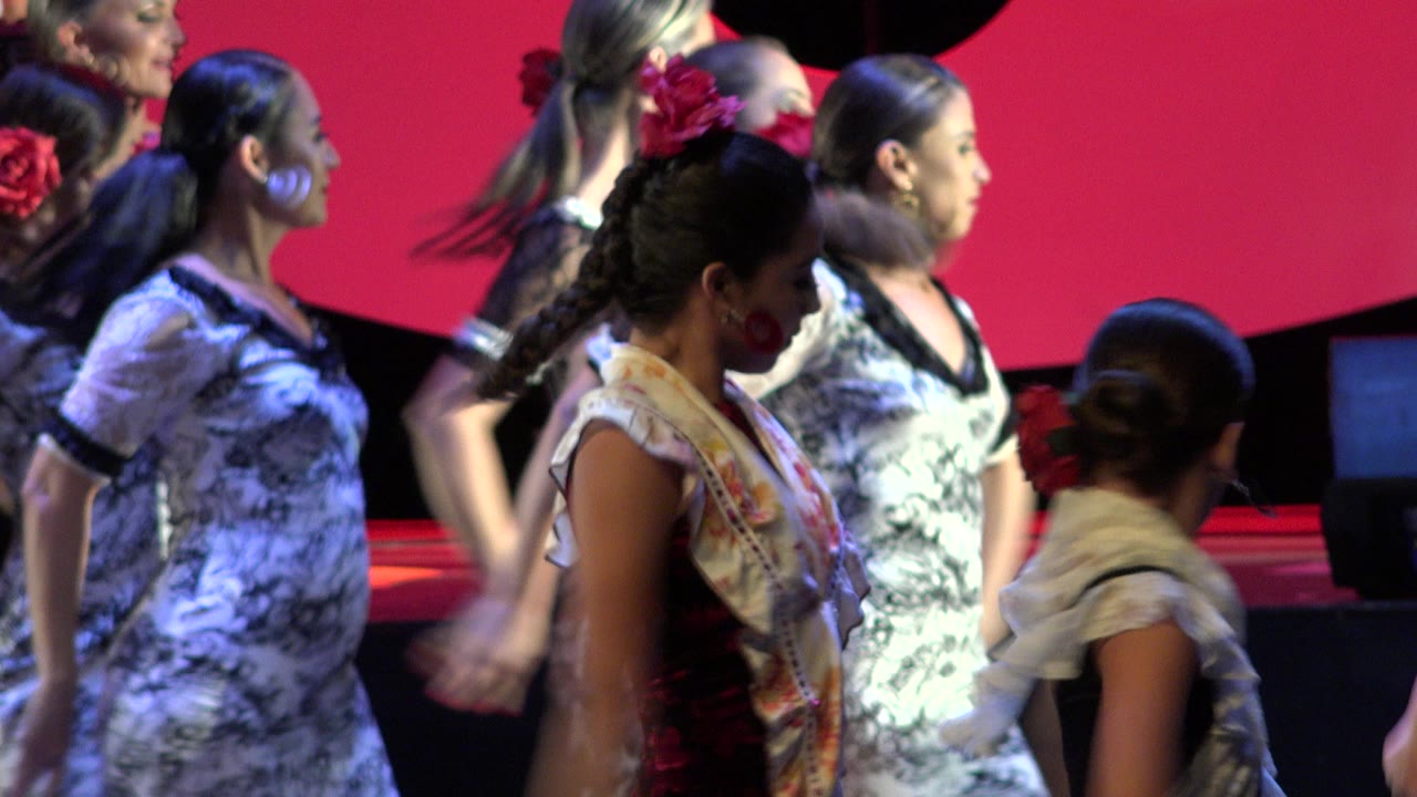 Dancers in patterned dresses remain in close stage formation in Cabo San Lucas, Mexico, with one young performer crossing the foreground and adult dancers behind her.