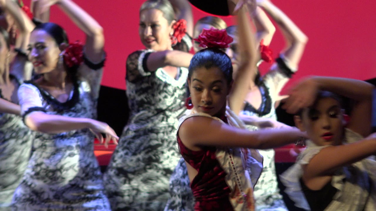 Women dancers perform close together on a Cabo San Lucas stage, with red hair flowers, patterned dresses, raised arms, and a red backdrop behind them.