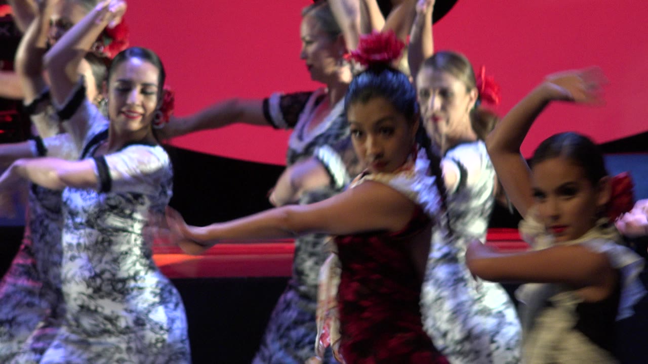 Dancers extend their arms across a stage with red backdrop graphics.