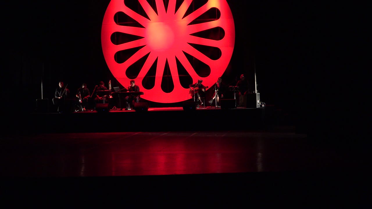At 0:01, the red wheel backdrop remains centered above the dark stage while the performers and equipment stay low in the frame.