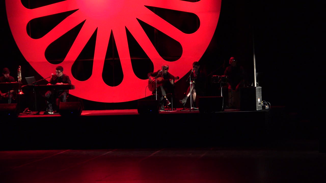 Wide view of a Cabo San Lucas concert stage with several seated musicians, black speakers, microphone stands, and a large red circular backdrop above them.