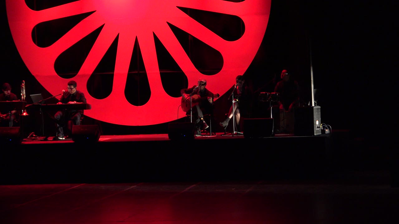 Cabo San Lucas concert stage with the guitarist near center-right, another musician at a keyboard on the left, and the red backdrop dominating the upper frame.