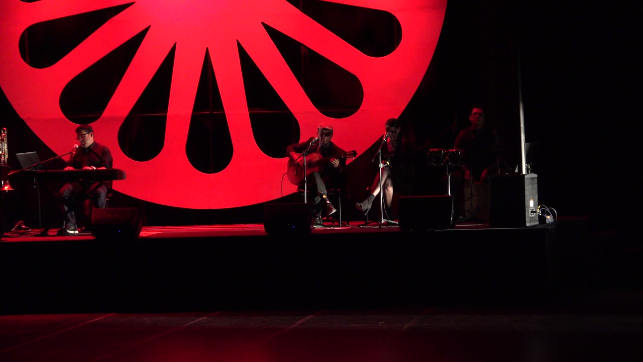 Guitarist and band seated on a dark stage with red backdrop.