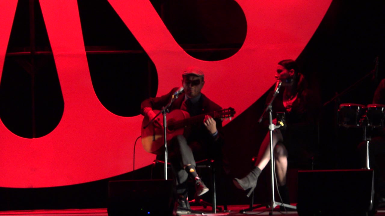 Seated guitarist sings into a microphone on a red-lit stage.
