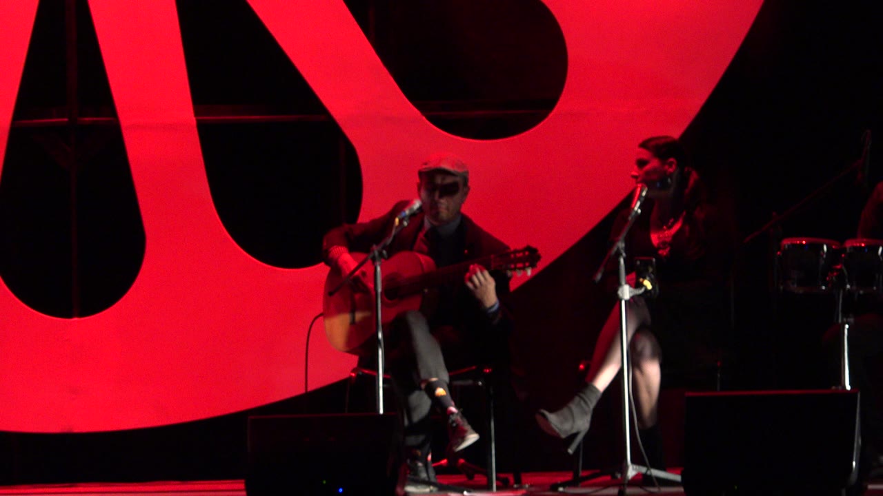 One second in, the Cabo San Lucas performance remains framed wide enough to show the guitarist, the seated woman, two microphone stands, and the oversized red-and-black backdrop.