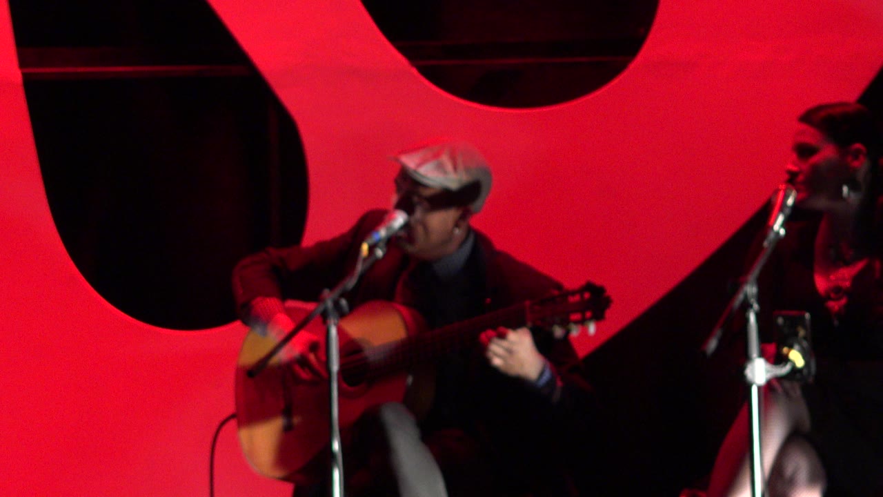 Man in hat playing acoustic guitar beside a woman at microphones on a red-lit stage.