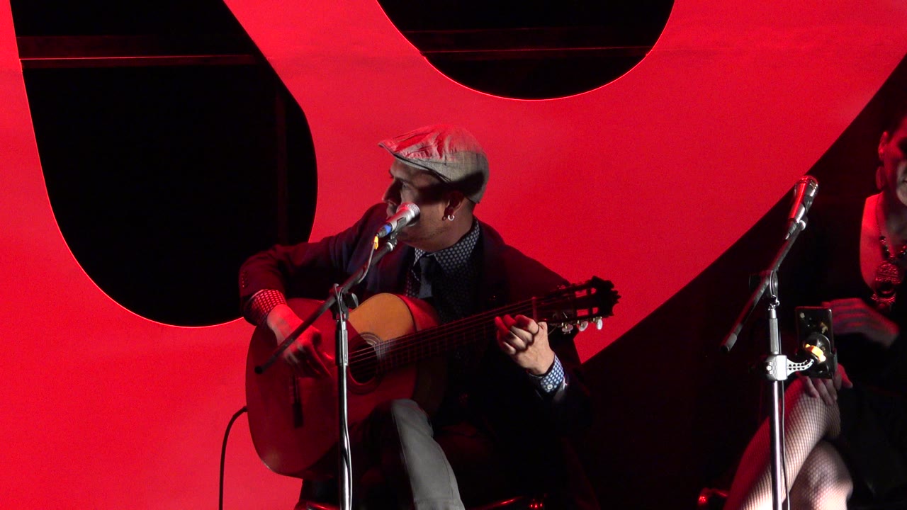 Man in a cap plays acoustic guitar under red stage light.