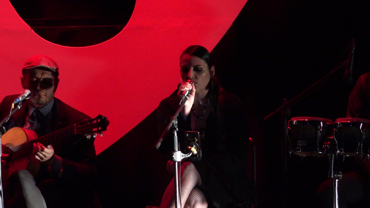 At 0:01 in Cabo San Lucas, Mexico, the singer remains close to the microphone with her hand raised near it as the guitarist continues playing in the red-lit stage frame.
