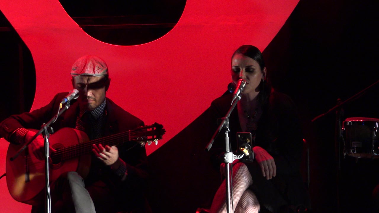 At 0:00 in Cabo San Lucas, Mexico, a seated man plays acoustic guitar while a woman sits beside a microphone against a red-lit stage backdrop.