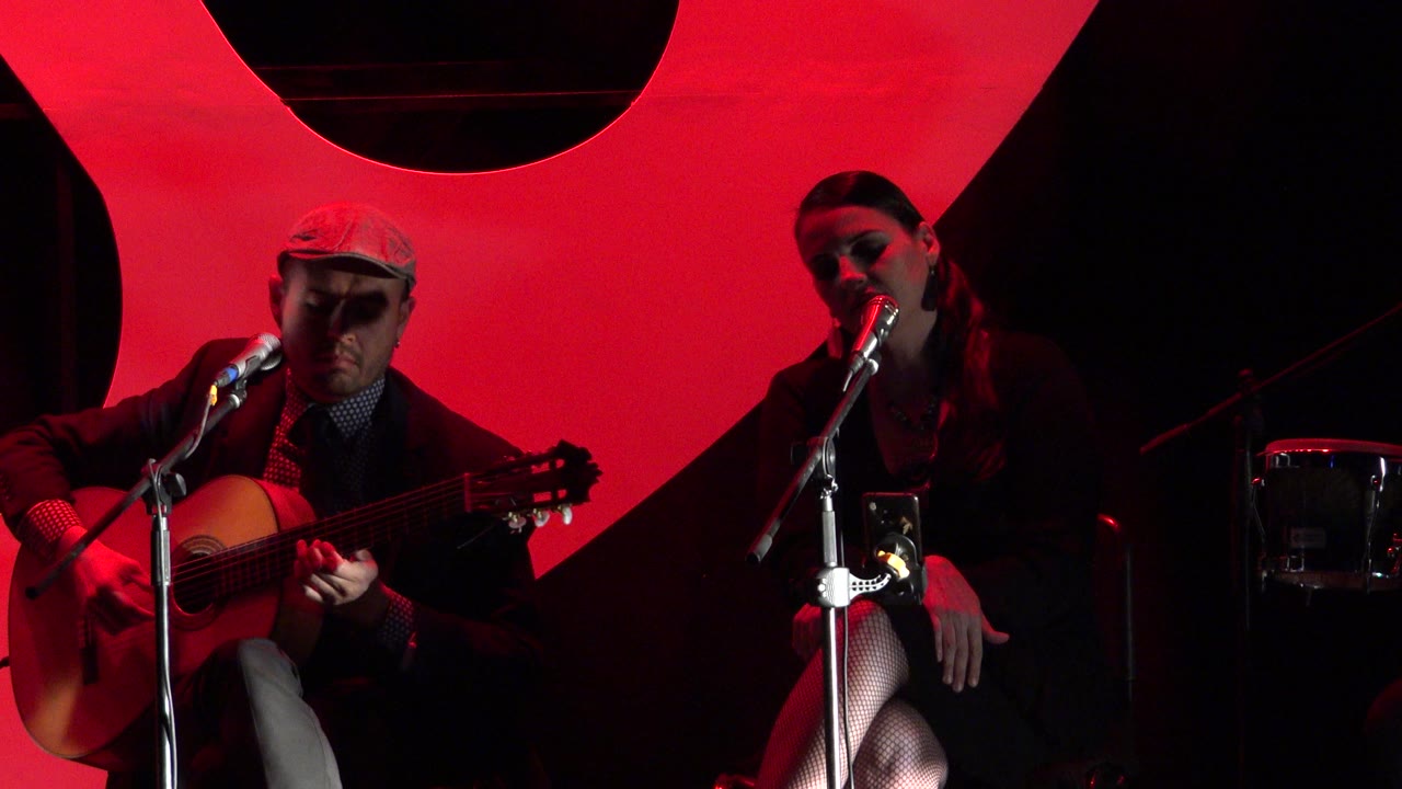 In Cabo San Lucas, Mexico, the guitarist looks down toward the instrument as the woman sits near the microphone in front of the red backdrop.