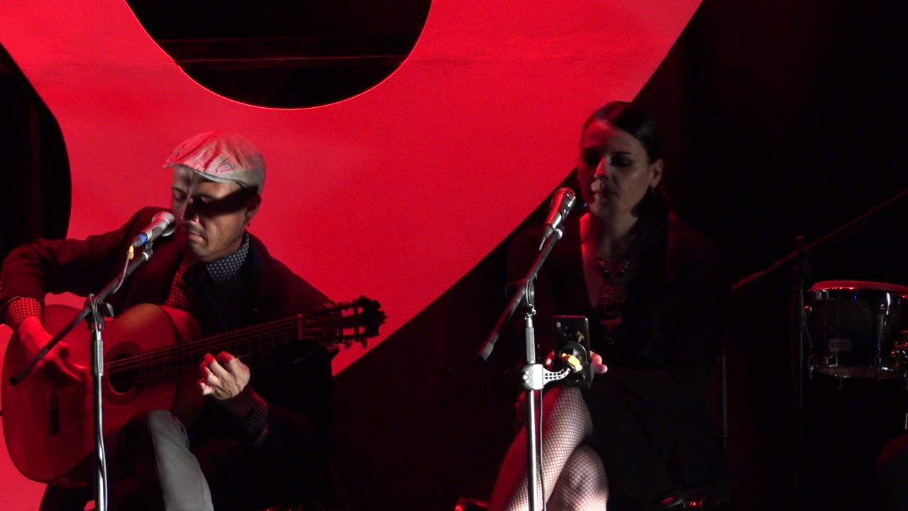 In Cabo San Lucas, Mexico, a seated guitarist plays beside a seated female singer at microphones under strong red stage lighting.