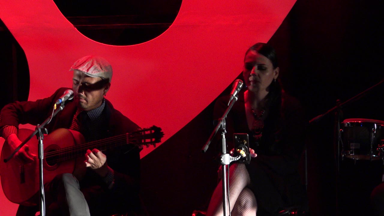 The guitarist remains centered with his instrument while the female vocalist sits at frame right near a microphone in the red-lit Cabo San Lucas stage scene.