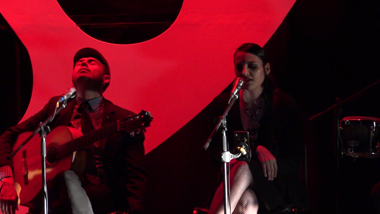 Two musicians on a red-lit stage with microphones and an acoustic guitar.