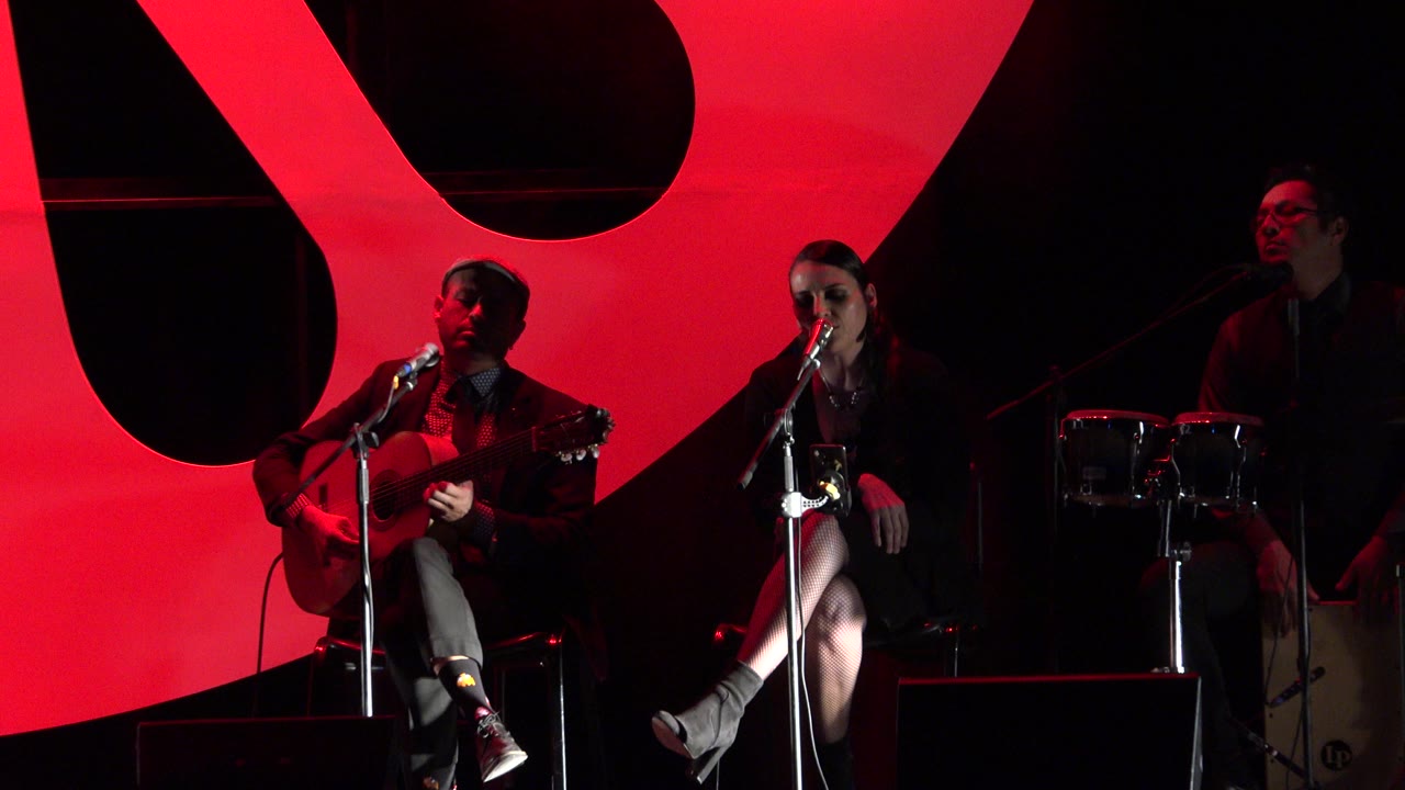 Musicians seated on a dark stage before a red backdrop.