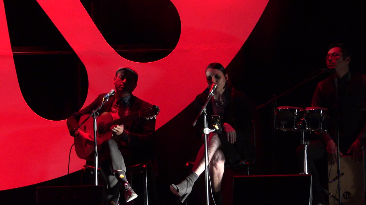 A guitarist and vocalist sit at microphones in Cabo San Lucas, Mexico, with a percussion setup partly visible against the red stage background.