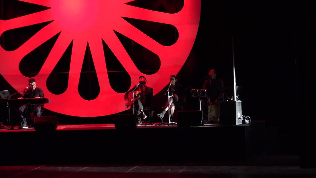 The Cabo San Lucas stage remains in a wide view, with a keyboard player near center, string players to the right, and the red radial circle glowing behind them.