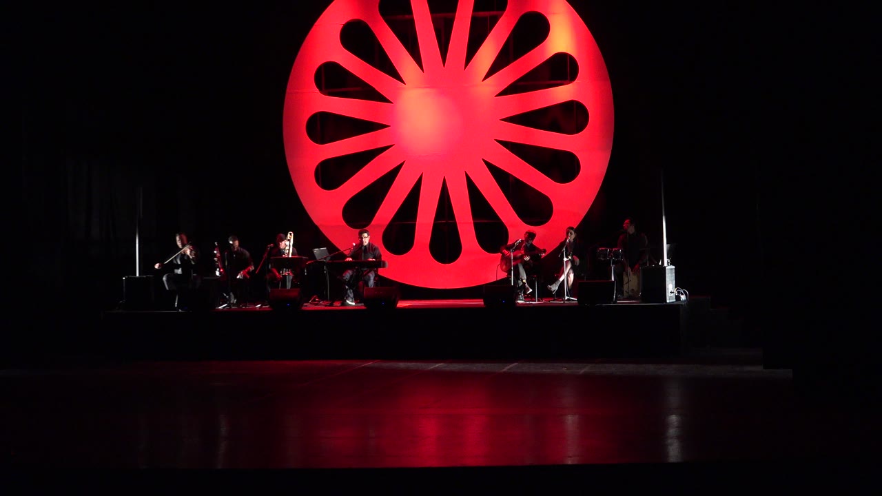 Wide stage with musicians under a red starburst backdrop.