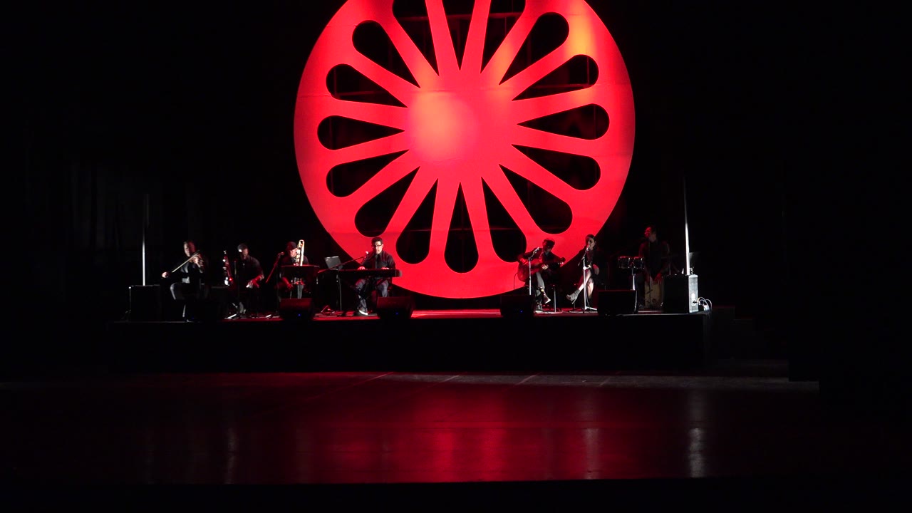 A small ensemble remains seated across the lower stage in Cabo San Lucas while the red starburst backdrop glows behind them.
