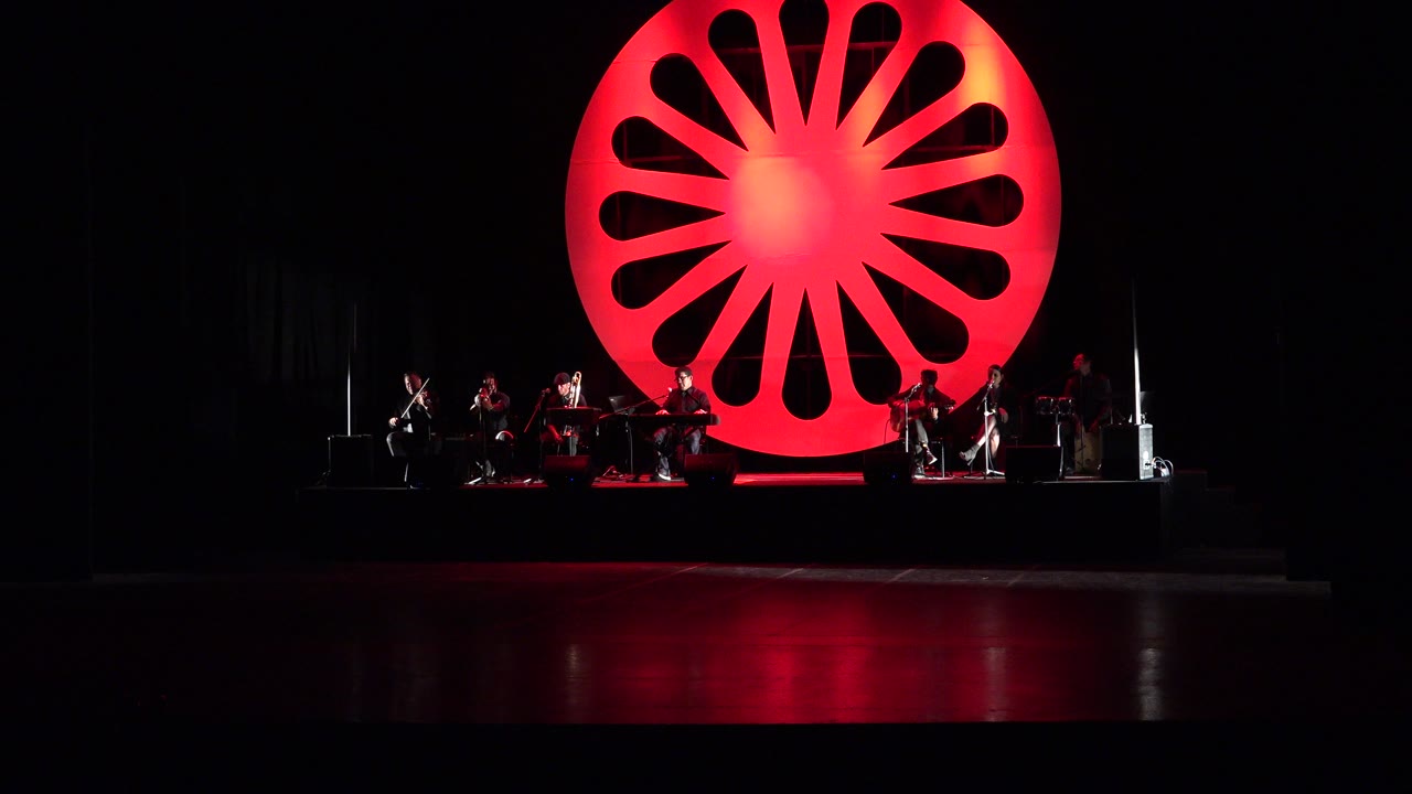 Wide dark stage with orchestra beneath a red circular backdrop.