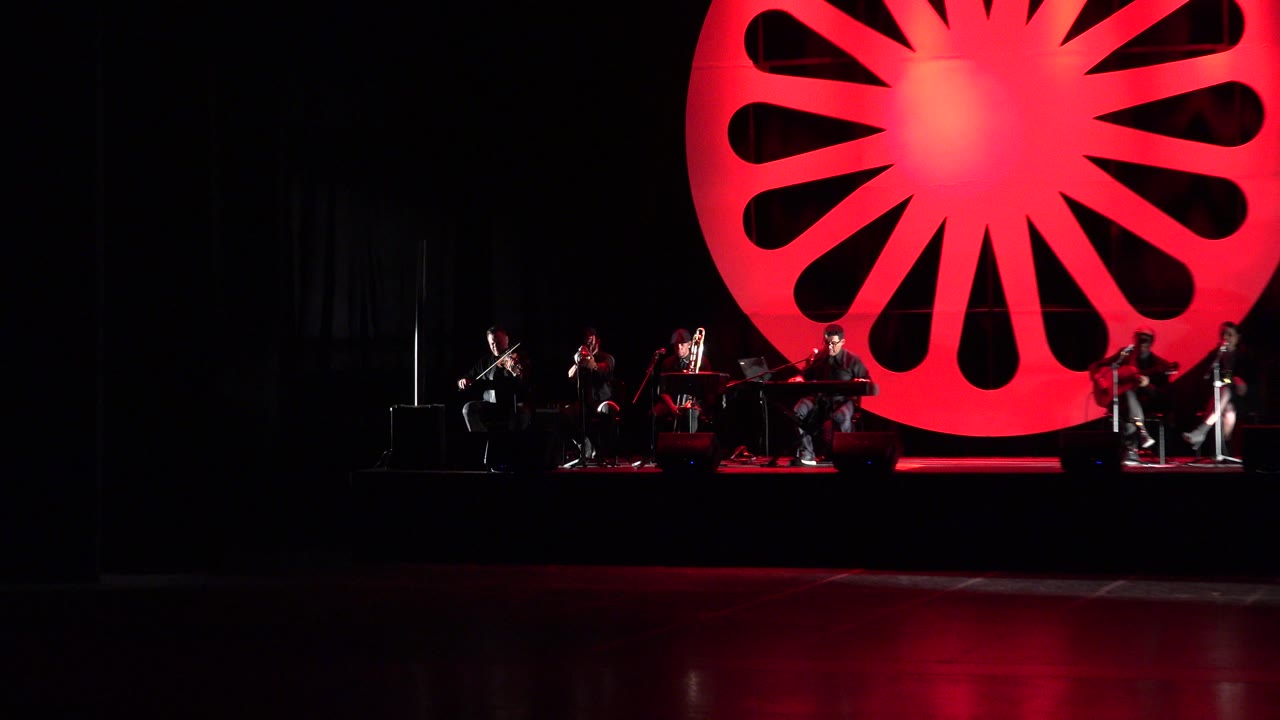 Wide dark stage with musicians and a red circular backdrop.
